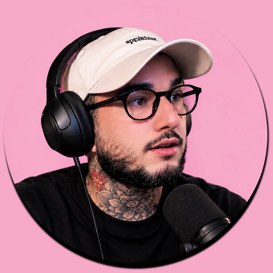 Hugo Pontes de boné claro, óculos e fone, falando ao microfone em um estúdio de fundo rosa.